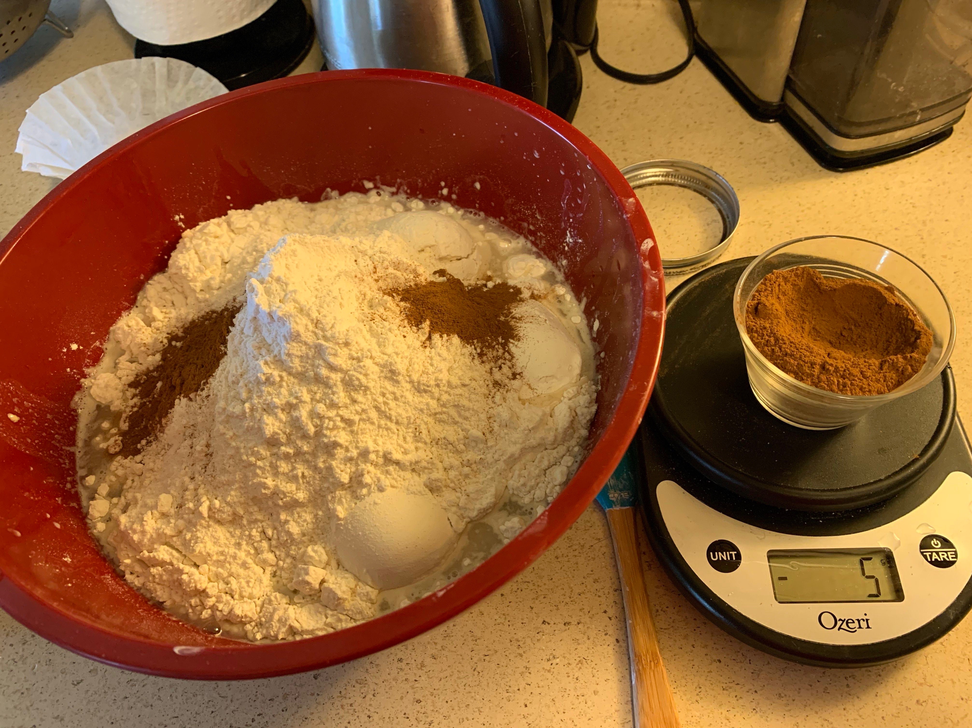 bowl of flour with two teaspoons of cinnamon added next to a kitchen scale reading -5g with a cup of cinnamon on top