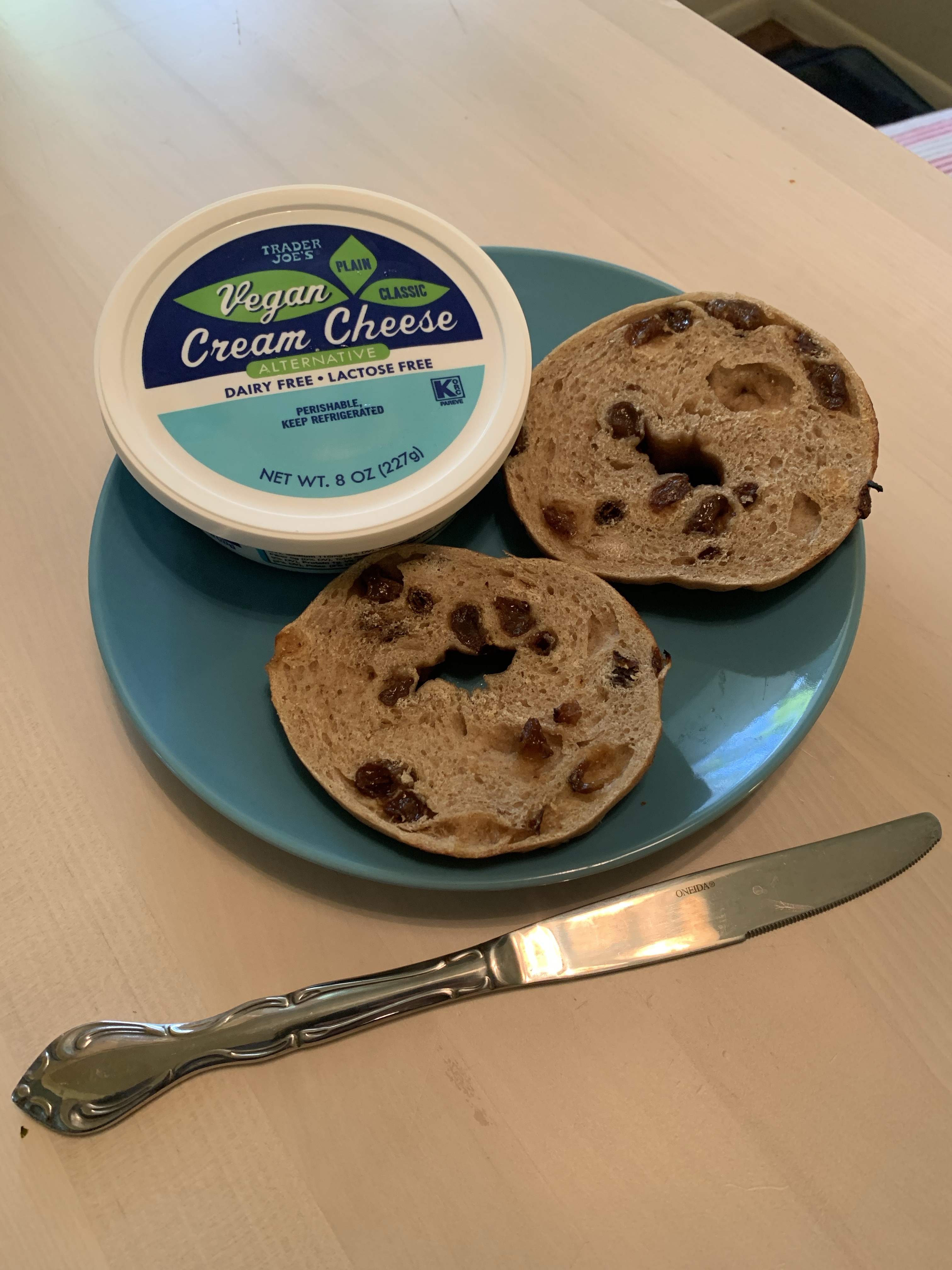 blue plate with container of vegan cream cheese and sliced open bagel on a table next to a butter knife