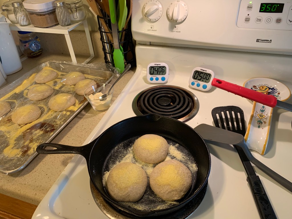 On the right is the counter with a baking pan. Is has several balls of dough covered in cornmeal and plastic wrap. Next to it on the counter is a small glass bowl filled with flour and a spoon. On the front left stove burner is a skillet with three dough balls and cornmeal. The dial is set to 3. Behind the top left burner are two digital timers reading 4:06 and 5:00. Next to the burners are two spatulas and a spoon rest. The oven is set to bake at 350°.