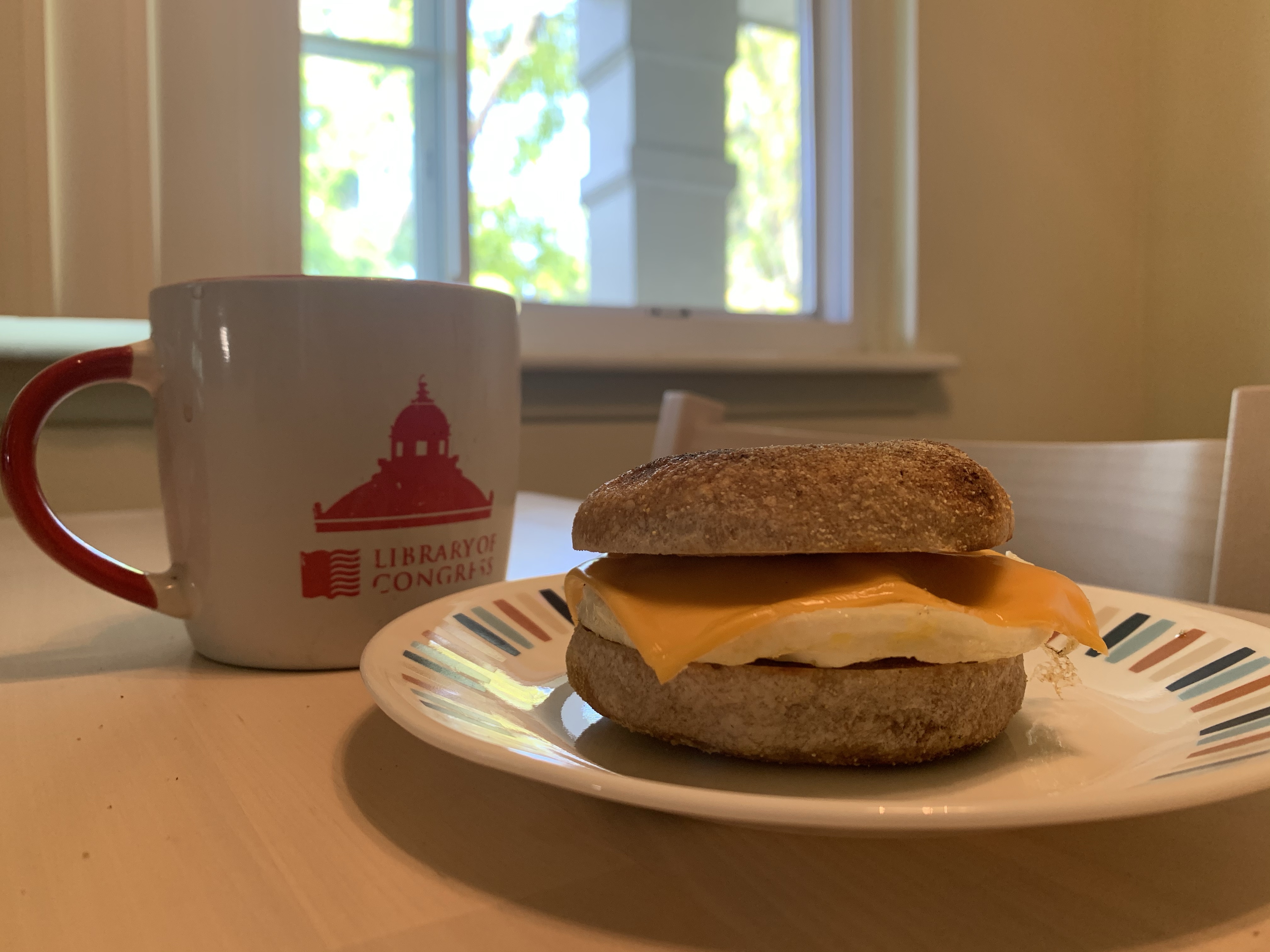 Sandwich of egg and chese on english muffin on plate by coffee mug. All on a table near an open window.