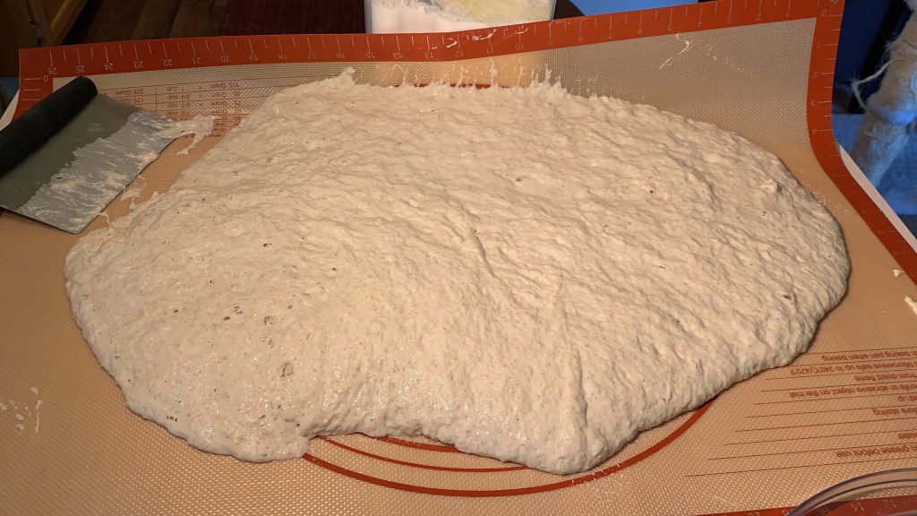 Rubber mat loaded with large mass of dough. Fold of one edge of mat is propped up with a flour container.