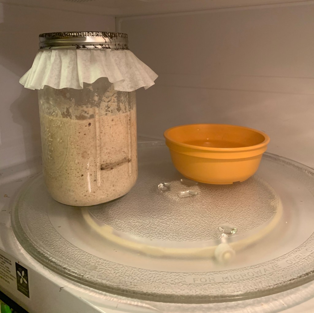 Inside of a microwave, there is a small, plastic, orange bowl of water and a mason jar. The mason jar is 2/3 full of bubbly starter, risen well past a dark line showing starting height. It is topped with a coffee filter and metal ring.