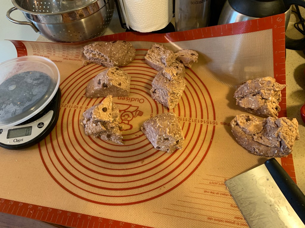 Rubber mat on kitchen counter. From left to right is a kitchen scale with a clear plastic lid, six hunks of cut dough, two larger hunks of cut dough, and a metal bench scraper.
