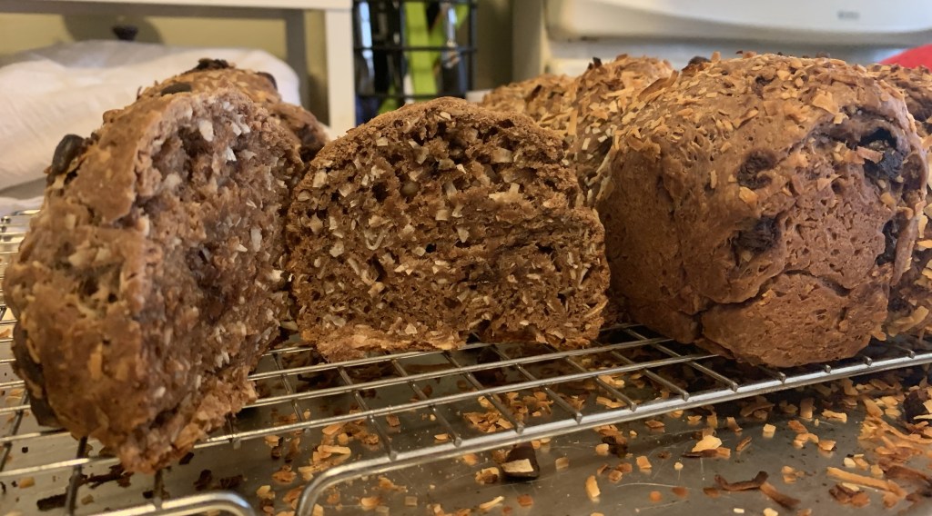 Lots of baked choco pan de coco on a wire rack. One piece is cut open and showing the crumb full of coconut bits.