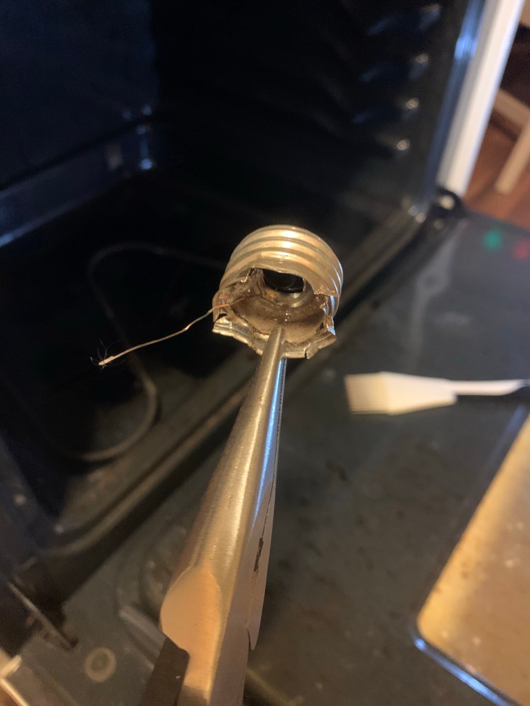 Pliers hold a mangled metal lightbulb socket in front of an open oven.