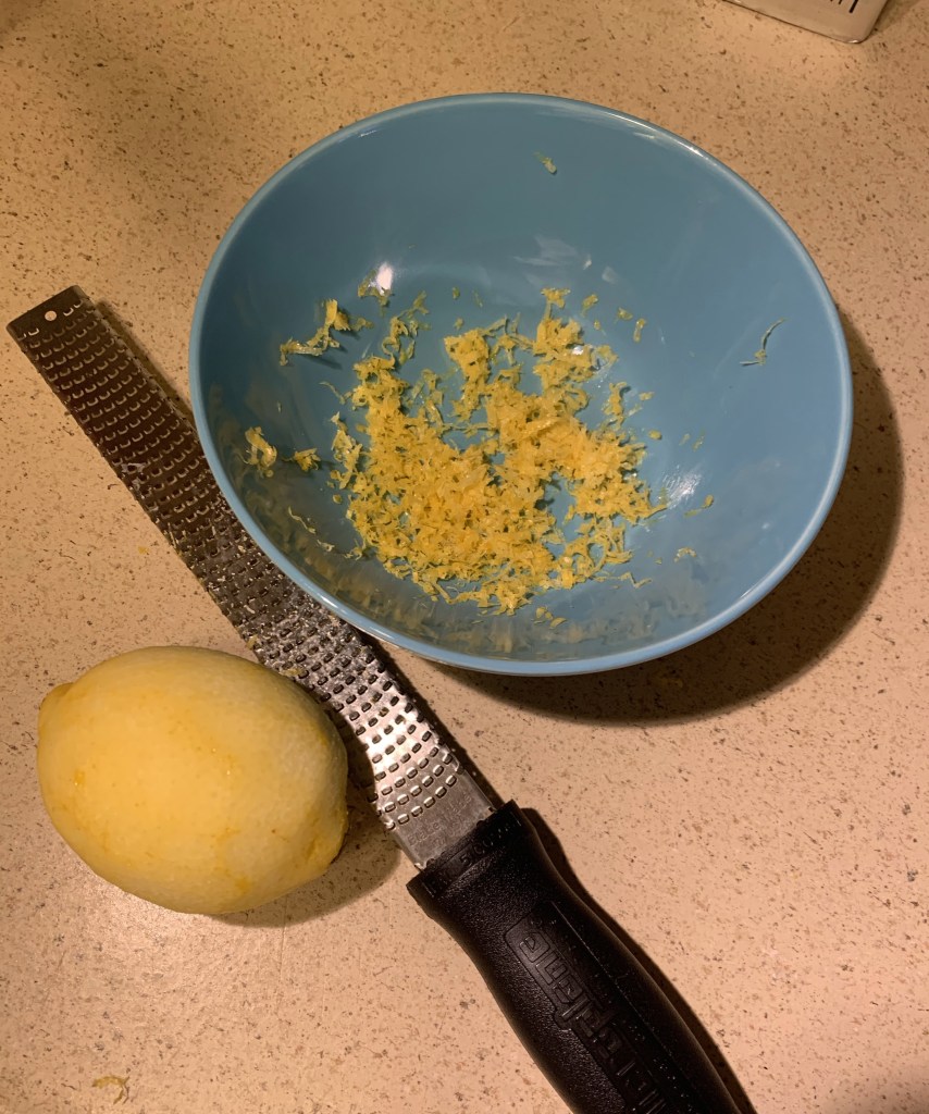 Blue bowl with fresh lemon zest on counter next to a microplane grater and a grated whole lemon.