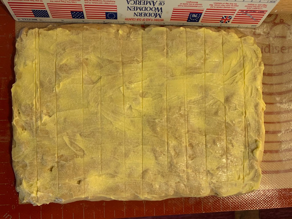 Rectangle of dough, smeared with butter and sugar, and showing eleven roughly parallel verticle lines. Beneath is a rubber mat with an embebbed ruler. Above is an actual plastic ruler with six "historic flags of our country," i.e., six versions of the U.S. flag. It reads "Modern Woodmen of America."