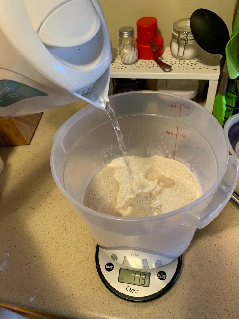 Water is pouring from a kettle into a clear container with flour on top of a kitchen scale reading 113g.