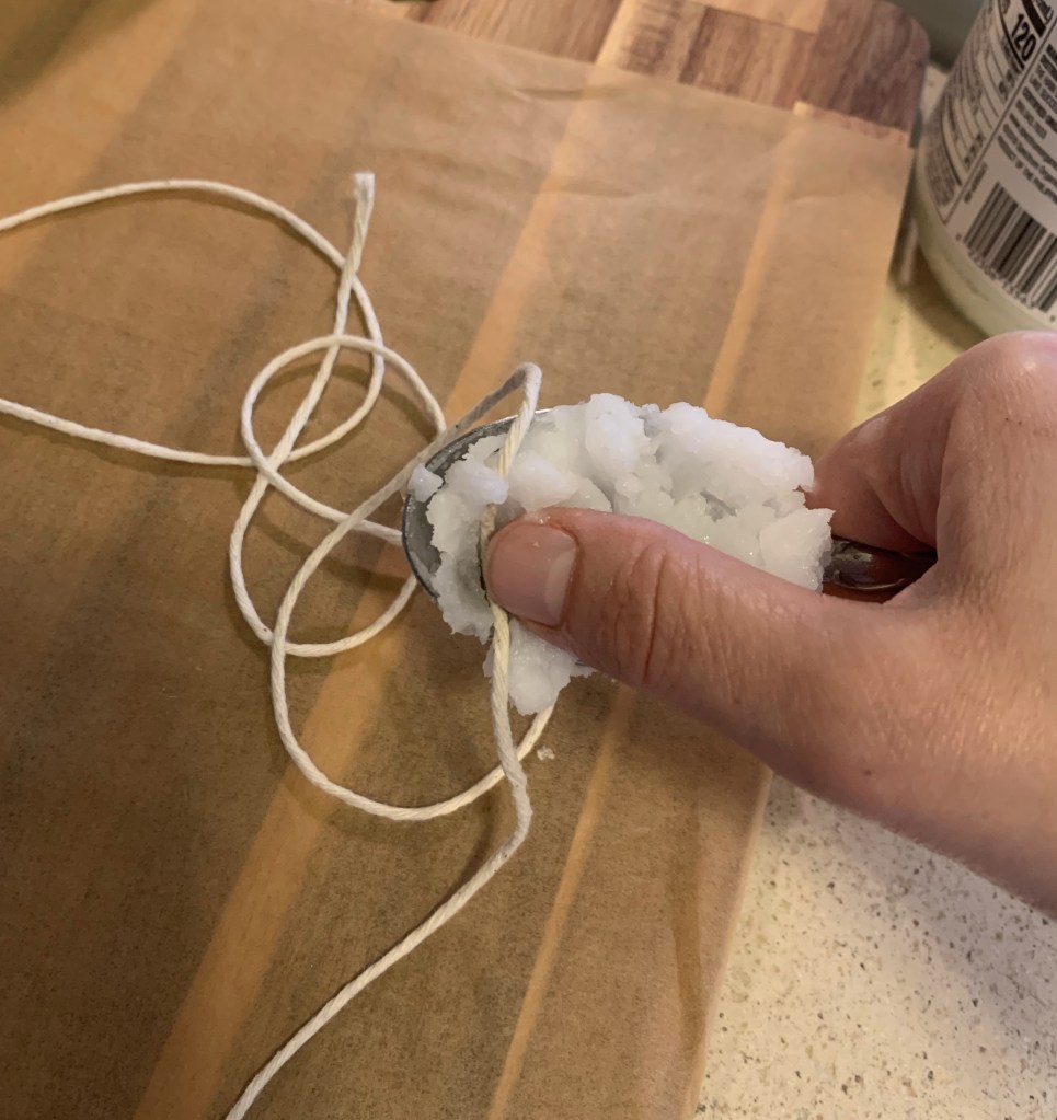 Hand holding a spoonful of solid coconut oil. The thumb is pressing a piece of twine into the oil. The rest of the twine is on a piece of parchment paper on a wooden cutting board.